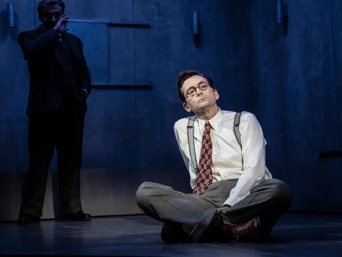 Photograph of David Tennant wearing a dress shirt, tie and slacks, sitting cross legged on a theater floor