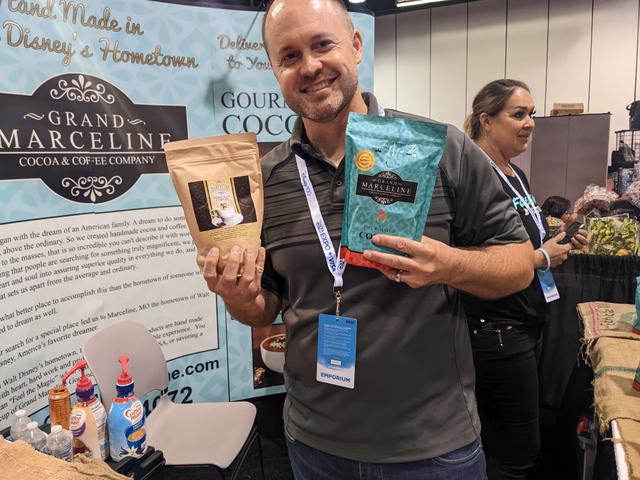 Joe Peck holding a bag of coffee and a bag of cocoa at the Grand Marceline booth