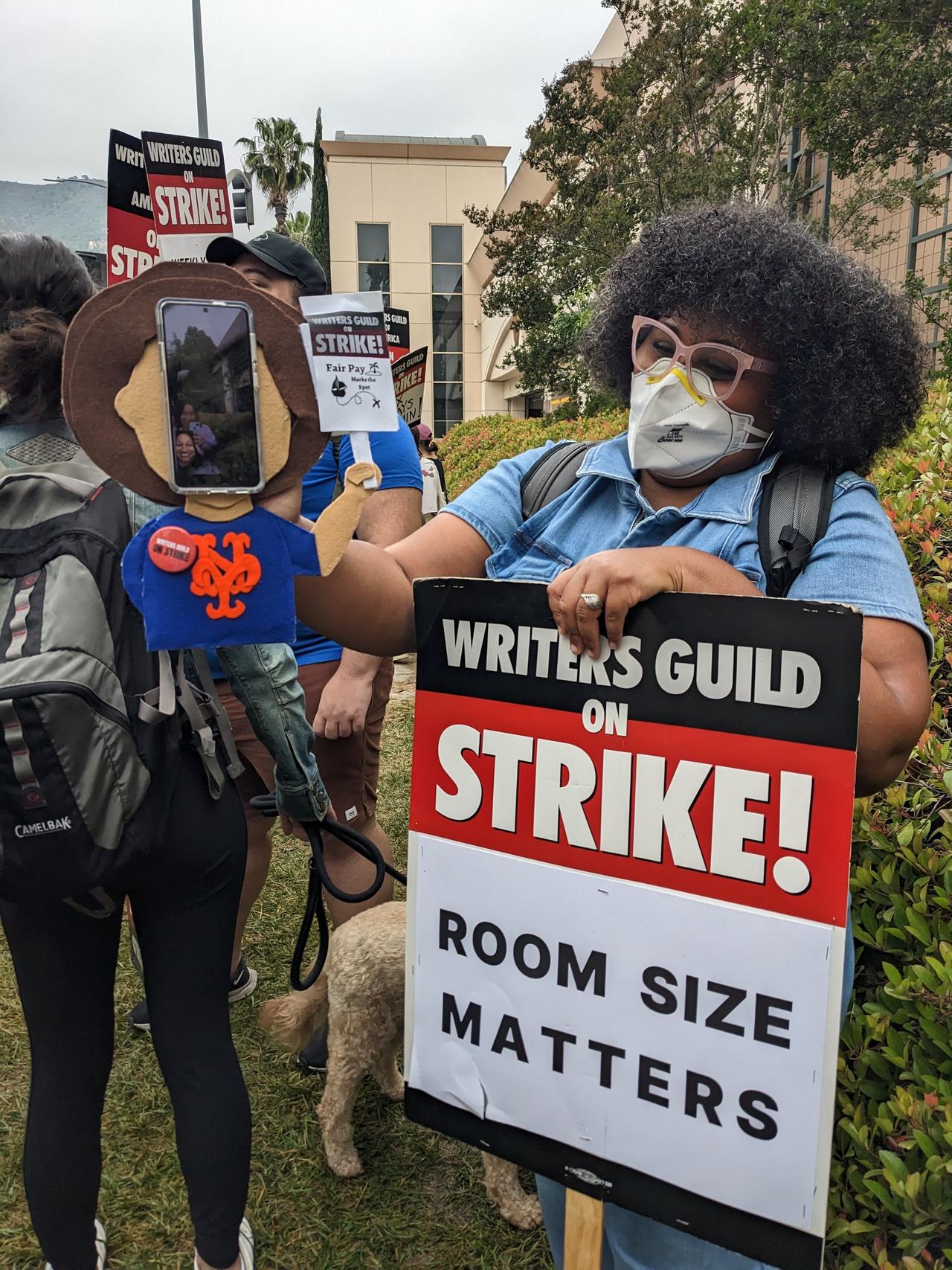 Photograph of a woman resting her hand on a picket sign and holding a felt puppet of a writer which holds a phone