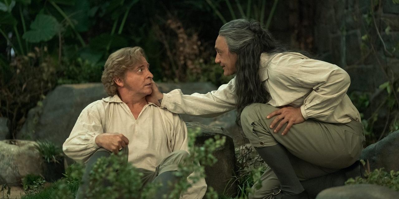 Our Flag Means Death HBO still with Rhys Darby as Stede Bonnet sat on the ground on the left, looking at Taika Waititi as Blackbeard who is holding his cheek on the right,